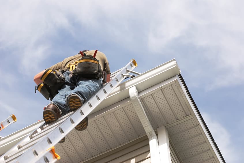 A handyman repairs his rain gutters. He is up a ladder, photo taken from ground looking up, low angle view. He wears a tool belt, sky and clouds, good copy space.